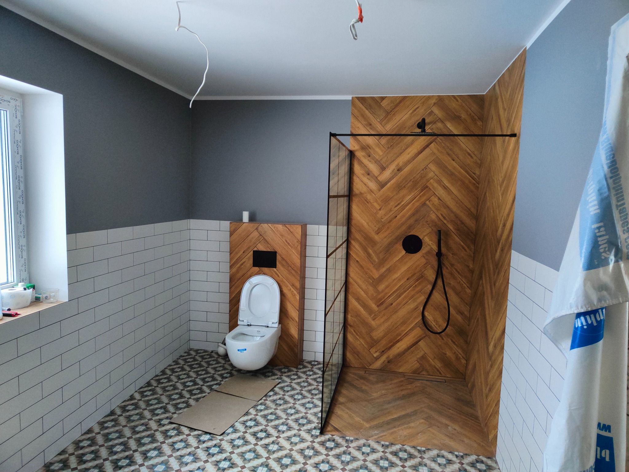 Nowoczesna łazienka w trakcie wykańczania: białe płytki na ścianach, wzorzysta podłoga, kabina prysznicowa z drewnopodobną ścianą w jodełkę, wisząca toaleta, widoczne kable elektryczne zwisające...