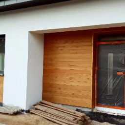 Fragment elewacji domu z drewnianą okładziną, widoczne okna z czarnymi ramami oraz drzwi wejściowe zabezpieczone folią ochronną, przygotowane do dalszych prac wykończeniowych.