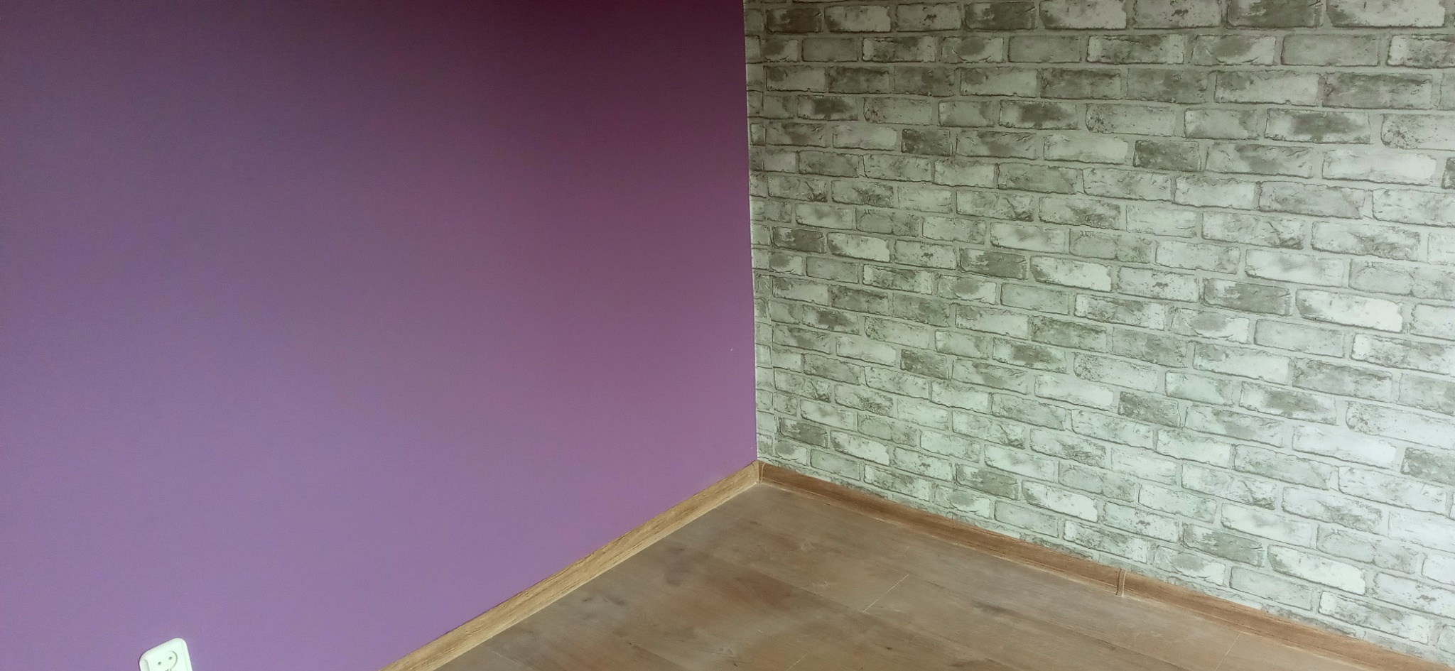 Różowa ściana sąsiadująca ze ścianą wykończoną tapetą imitującą białą cegłę w rogu pomieszczenia w Sieradzu, z widoczną podłogą z paneli.