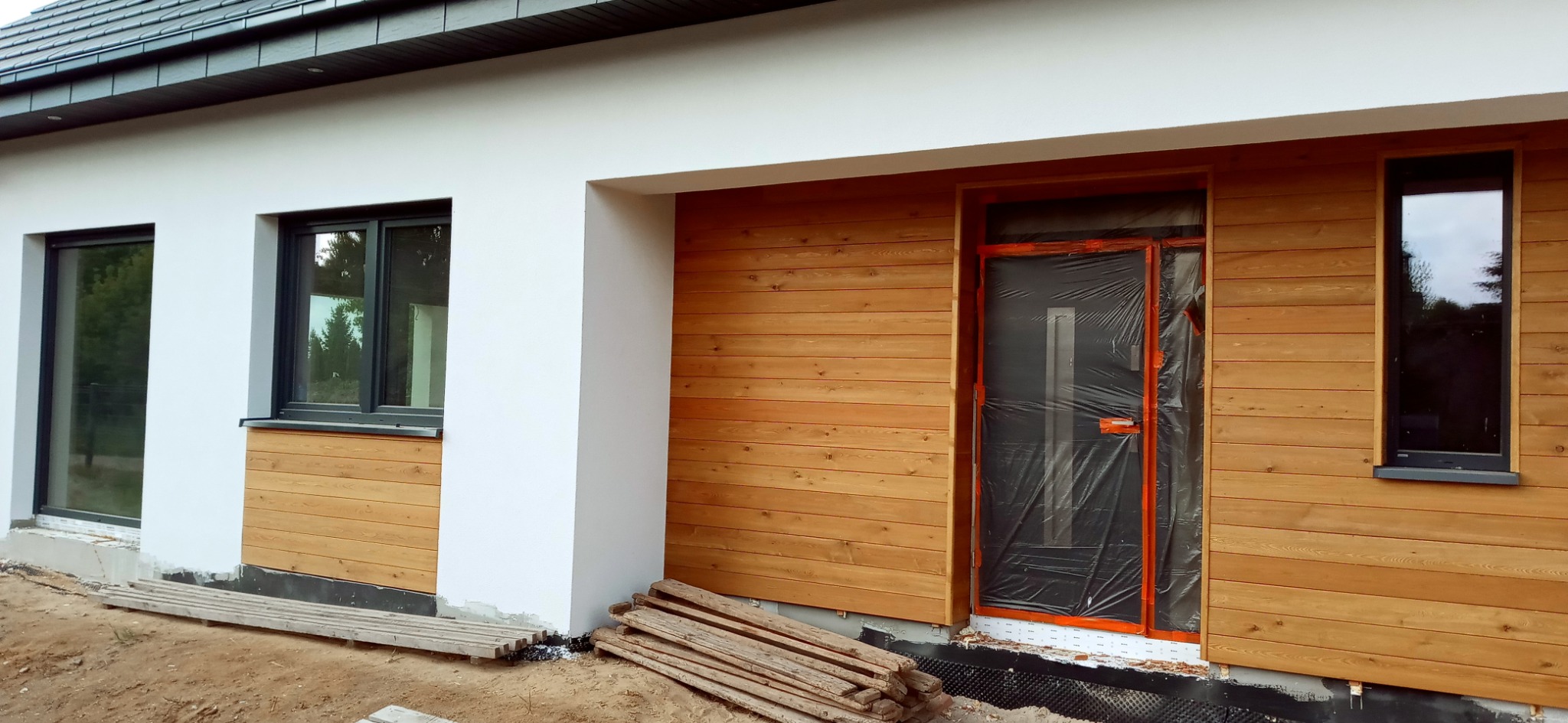 Fragment elewacji domu z drewnianą okładziną, widoczne okna z czarnymi ramami oraz drzwi wejściowe zabezpieczone folią ochronną, przygotowane do dalszych prac wykończeniowych.