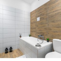 Adrian Król - Nowoczesna łazienka z białymi i drewnopodobnymi płytkami, czarnymi lampionami dekoracyjnymi na podłodze i minimalistycznymi akcentami roślinnymi.