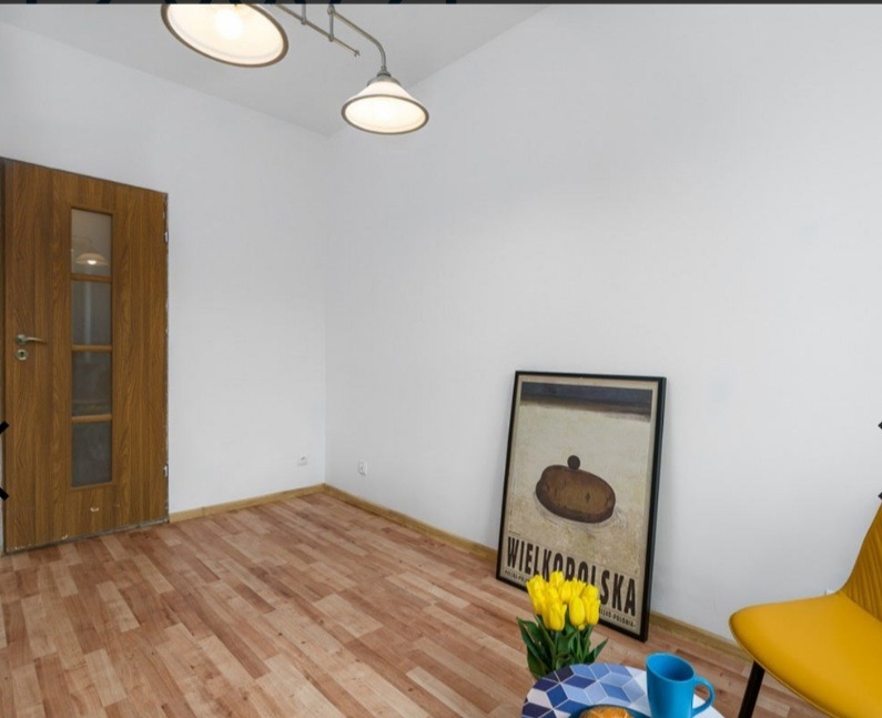 Pusty pokój po odświeżeniu z panelami podłogowymi, białymi ścianami, drewnianymi drzwiami z szybkami, lampą na suficie i dekoracyjnym plakatem Wielkopolska na ścianie.