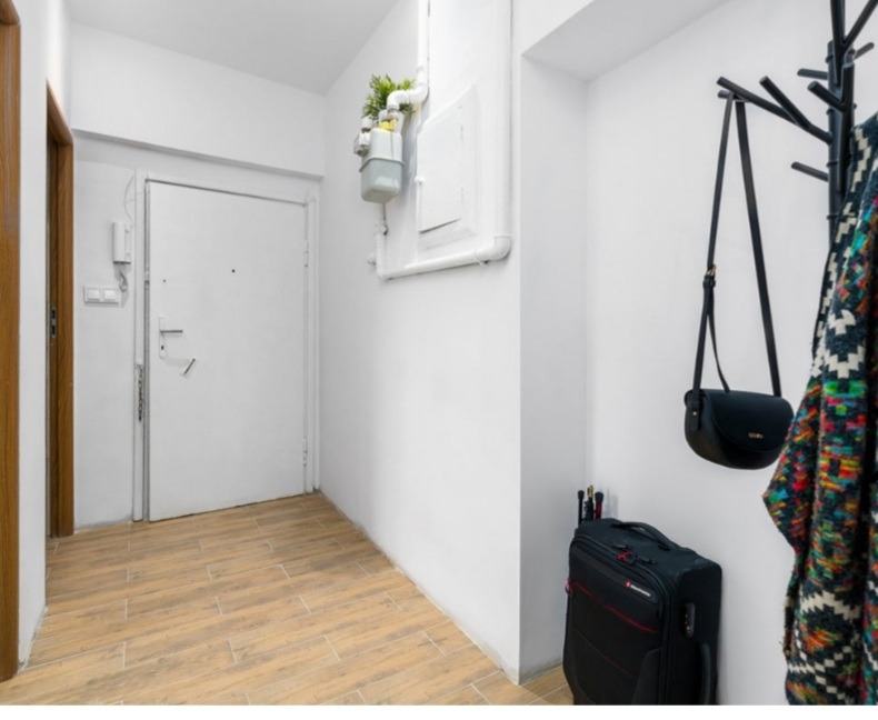 Przedpokój z jasnymi ścianami i drewnopodobną podłogą, widoczny biały licznik gazowy z rośliną na rurze, czarny wieszak z torbą i kolorową narzutą, czarna walizka z akcesoriami.