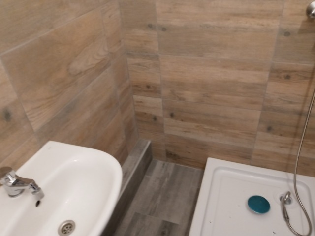 Umywalka narożna z chromowaną baterią i kabina prysznicowa z niebieskim korkiem odpływowym, wykończone płytkami imitującymi drewno w jasnych odcieniach beżu i brązu.