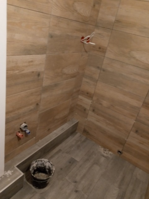 Ściana i podłoga w łazience wyłożone płytkami imitującymi drewno, widoczne wystające instalacje wodne i elektryczne oraz wiadro z zaprawą.