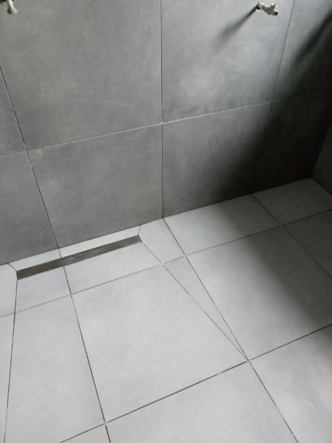 Ukośne ujęcie szarych, matowych płytek ściennych i podłogowych w łazience z liniowym odpływem podłogowym, widoczne niedokładności w fugowaniu.