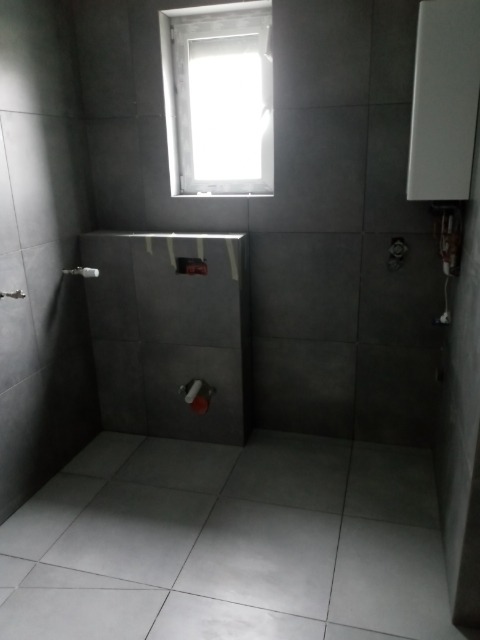 Szara łazienka w trakcie prac wykończeniowych: widoczne płytki na ścianach i podłodze, zabudowa podtynkowego WC ze stelażem, okno z białymi ramami i zamontowany biały piec gazowy na ścianie.