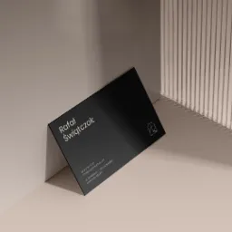 Elegancka, czarna wizytówka z minimalistycznym logo i danymi kontaktowymi Rafała Świątczaka, oparta o ścianę z dekoracyjnymi pionowymi żłobieniami, w stonowanej kolorystyce.