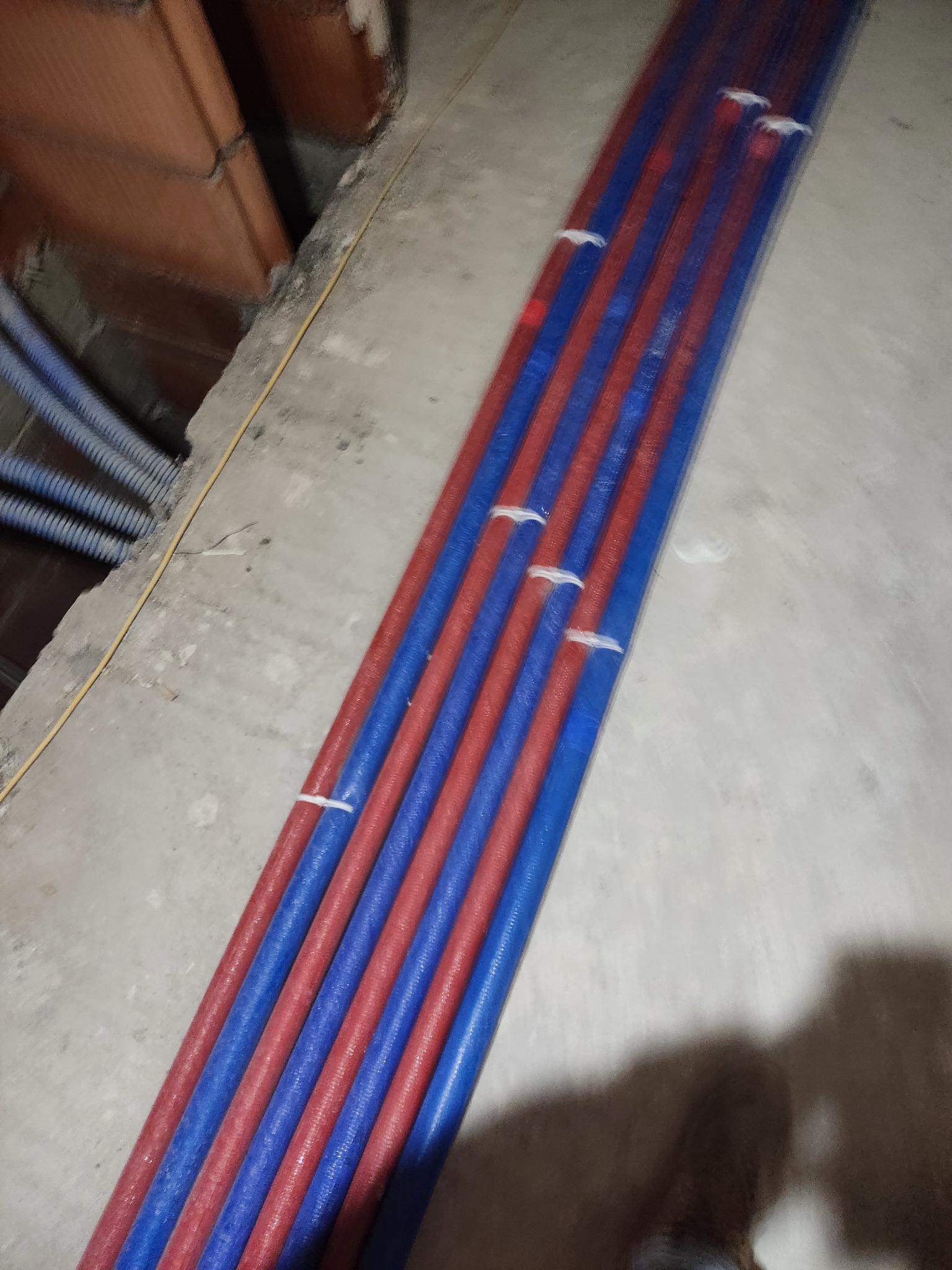 Równolegle ułożone rury instalacyjne w kolorach czerwonym i niebieskim, zabezpieczone białymi opaskami zaciskowymi, widoczne na surowym betonowym podłożu w trakcie prac instalacyjnych.