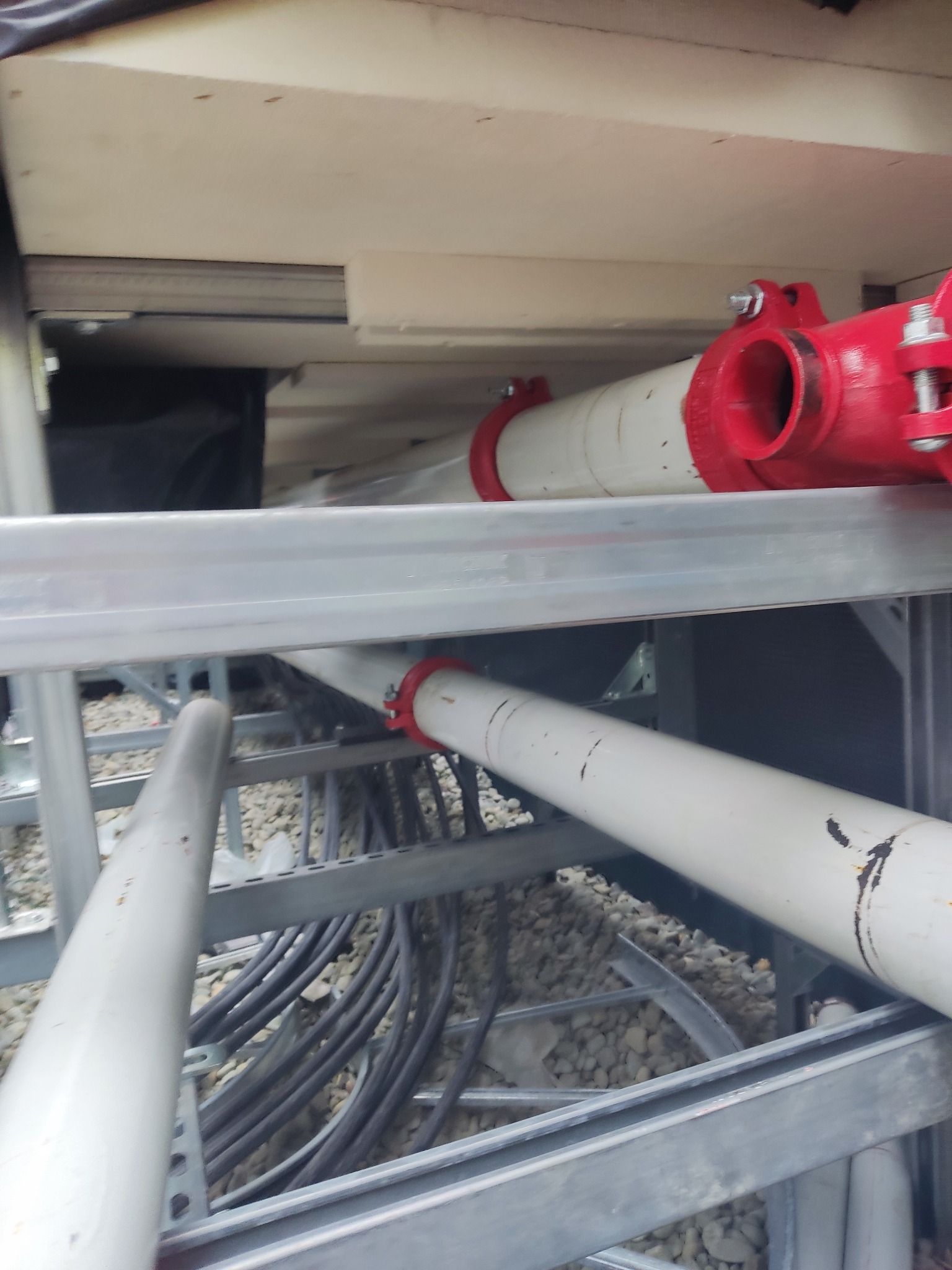 Białe rury z czerwonymi łącznikami zamontowane na metalowej konstrukcji wsporczej, widoczne kable i żwir na podłożu.