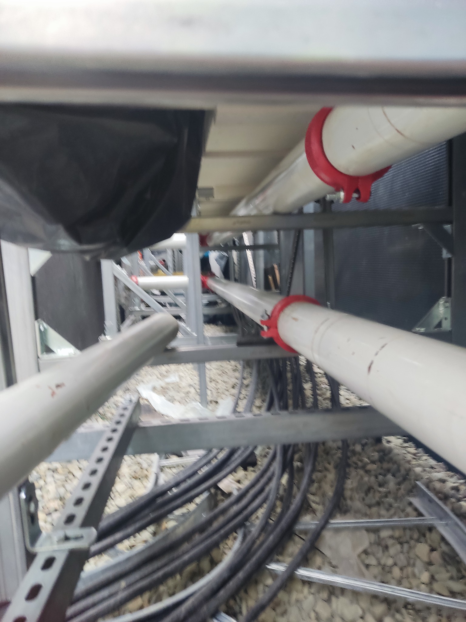 Widok z dołu na instalację rur i kabli w metalowej konstrukcji wsporczej z czerwonymi uchwytami, widoczne przewody i żwirowe podłoże.