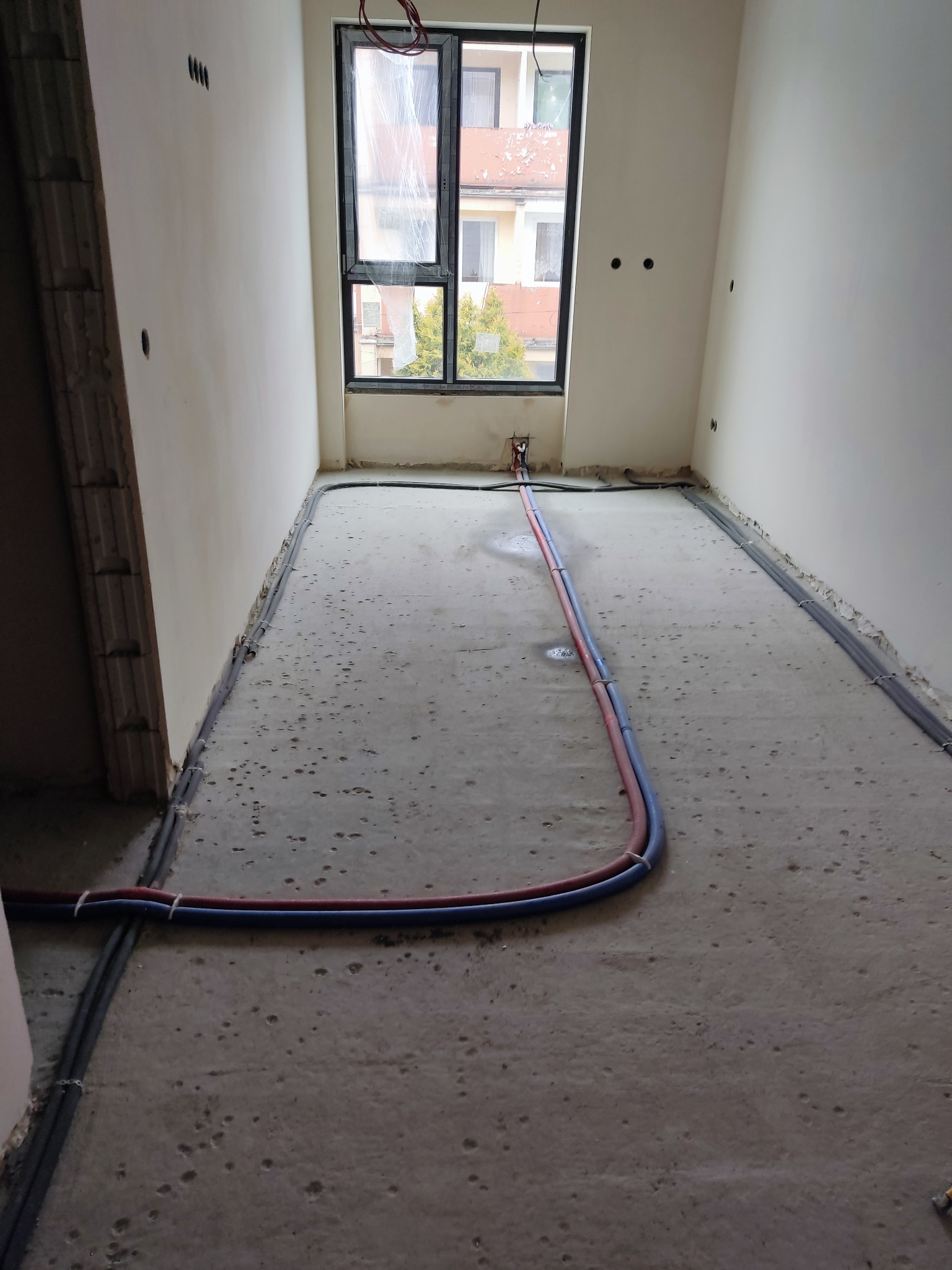 Surowy stan mieszkania w trakcie remontu: widoczne rury instalacji wodnej w kolorach niebieskim i czerwonym ułożone na betonowej posadzce, okno z widokiem na budynek, ściany przygotowane...