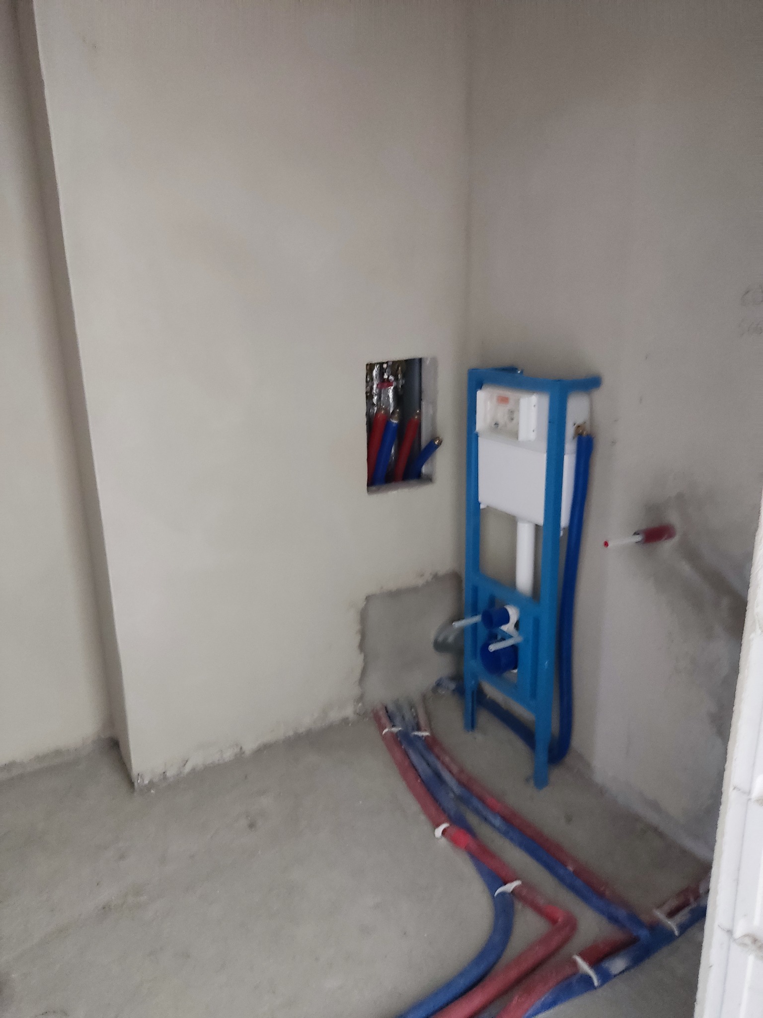 Surowe wnętrze łazienki w trakcie prac instalacyjnych: widoczne nieotynkowane ściany, rury wodne w kolorze niebieskim i czerwonym prowadzone po podłodze oraz stelaż podtynkowy WC z białym...