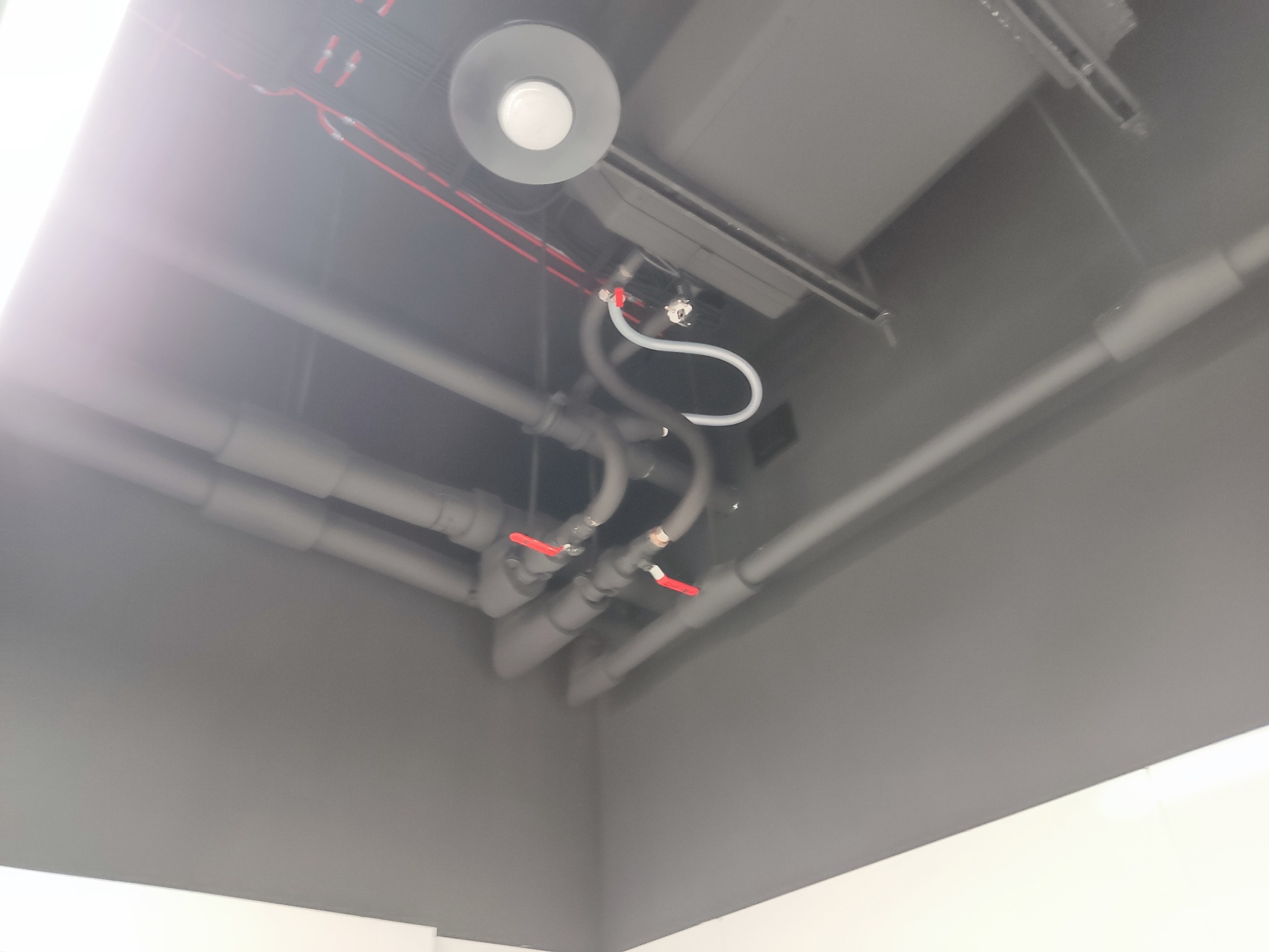 Szare rury instalacyjne z czerwonymi zaworami, biała rurka pod sufitem, widok z dołu, oświetlenie sufitowe.