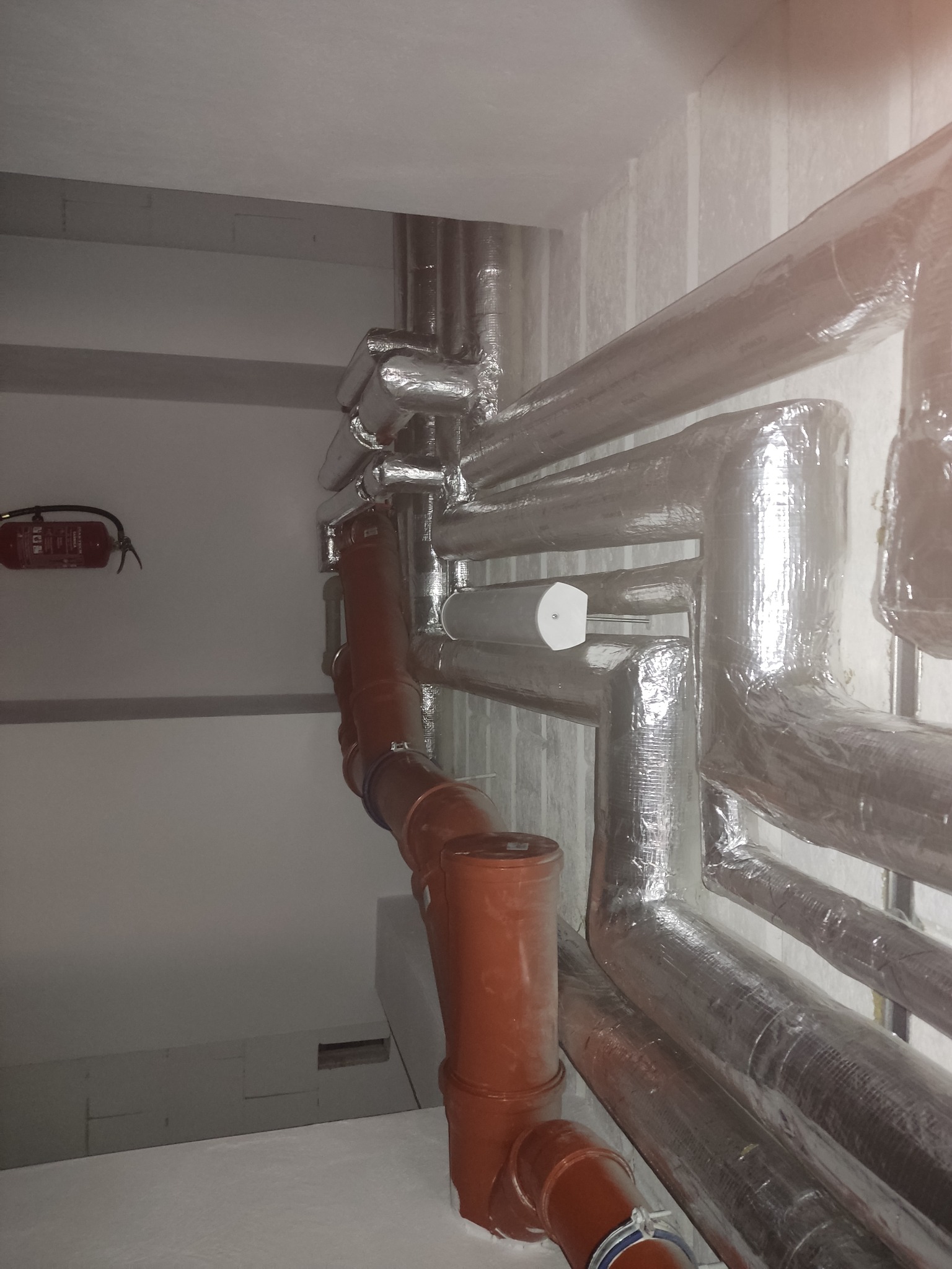 Rury kanalizacyjne z tworzywa w kolorze ceglastym oraz rury izolowane folią aluminiową, biegnące wzdłuż ściany w pomieszczeniu technicznym, z widoczną gaśnicą na ścianie.
