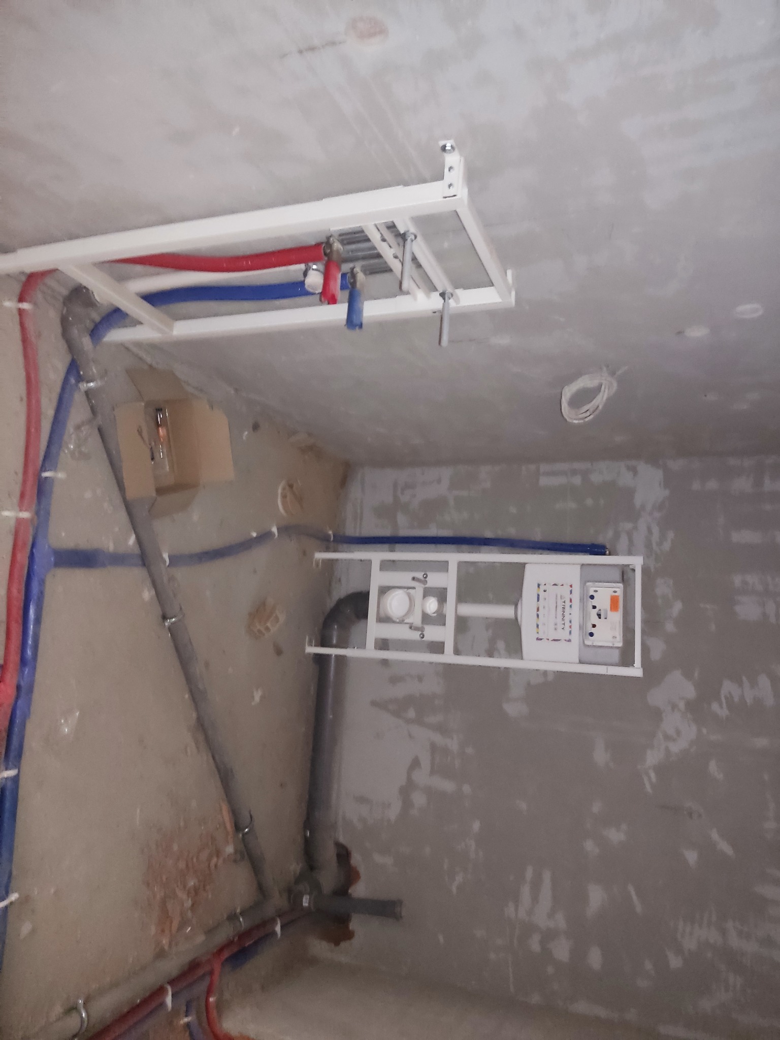 Surowe wnętrze łazienki w trakcie remontu, widoczne rury instalacji wodnej w kolorze czerwonym i niebieskim, stelaż podtynkowy WC z widoczną spłuczką i przyciskiem, oraz fragment podwieszanego...