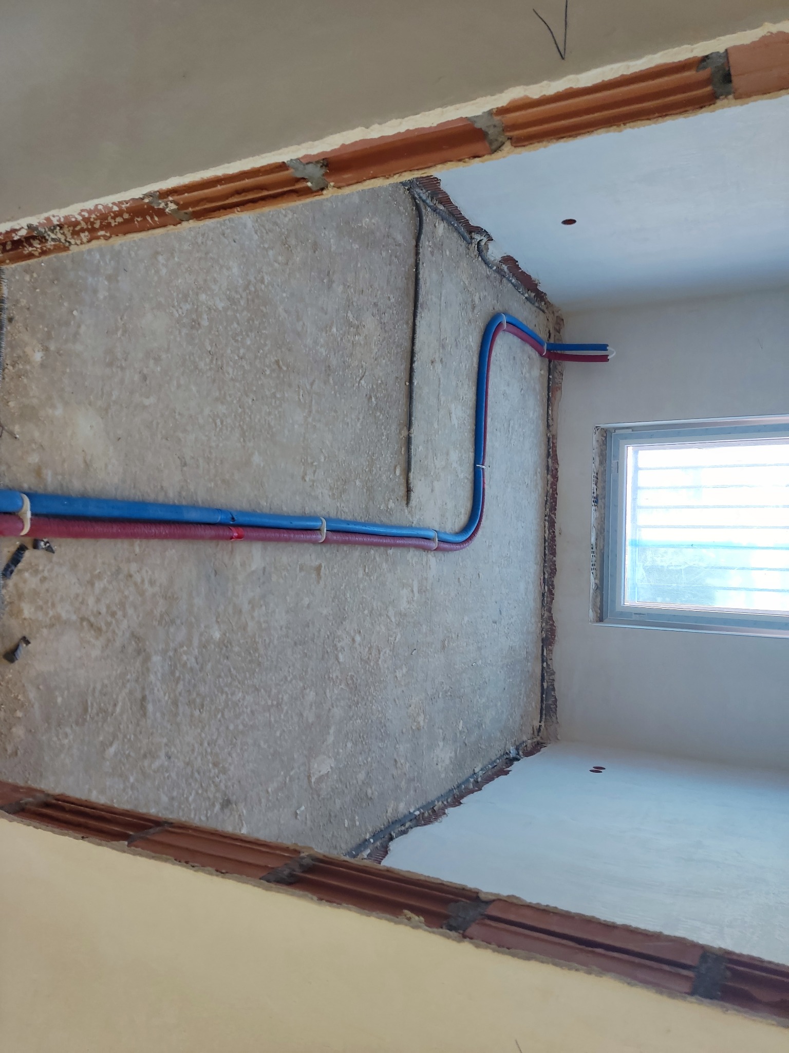 Wnętrze w trakcie remontu z widocznymi, nowymi rurami instalacji wodnej (czerwona i niebieska) poprowadzonymi wzdłuż ściany, surowa powierzchnia ściany z widocznymi śladami po kuciu, okno w tle.