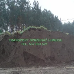 Góra ziemi ogrodowej (humusu) na placu budowy lub składowisku, widoczne ślady opon pojazdów, w tle las iglasty, na ziemi zielony napis informujący o transporcie i sprzedaży humusu oraz numer telefonu.