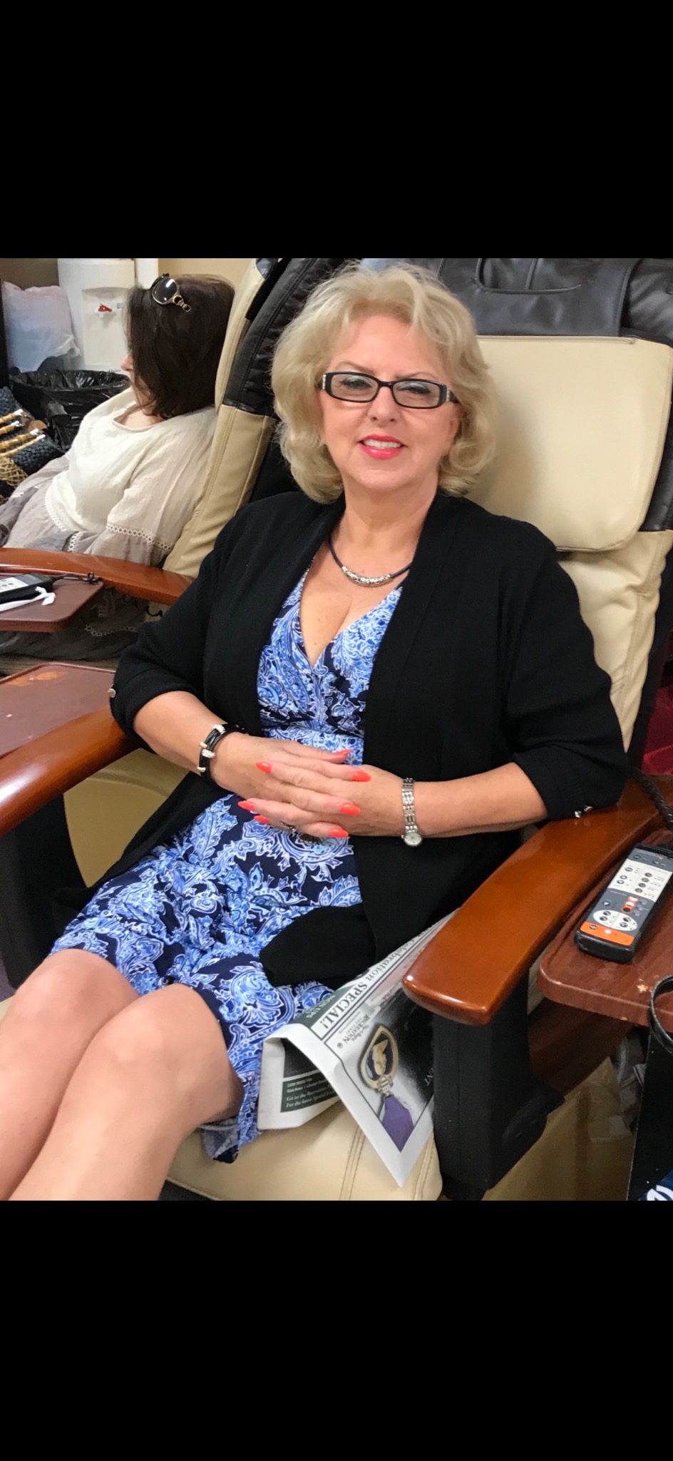 Kobieta w fotelu do pedicure, ubrana w niebieską sukienkę w paisley i czarny kardigan, z pomarańczowymi paznokciami i gazetą leżącą obok pilota.