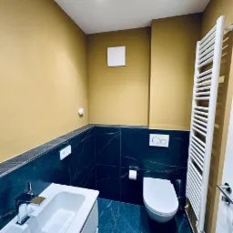 Nowoczesna łazienka z umywalką na szafce, toaletą i grzejnikiem drabinkowym na tle ścian w odcieniach złota i ciemnego marmuru.