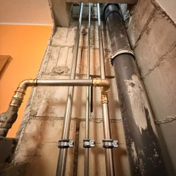HETVOLD Sp. z o.o. - Pionowy układ rur w surowym wnętrzu, z widocznymi mocowaniami i fragmentem instalacji kanalizacyjnej. Ściany z betonu komórkowego, pomarańczowy fragment ściany w tle.