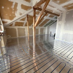 HetVold Sp. z o.o. - Instalacja ogrzewania podłogowego w surowym wnętrzu poddasza z drewnianą konstrukcją. Rury grzewcze ułożone na folii izolacyjnej, ściany przygotowane do malowania.