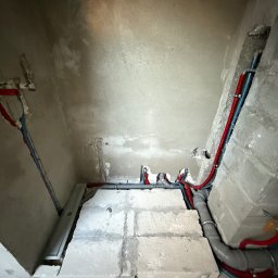 HETVOLD Sp. z o.o. - Surowy stan: Instalacja wodno-kanalizacyjna z rurami w kolorze czerwonym i niebieskim, szare rury kanalizacyjne, widoczne ściany w trakcie prac wykończeniowych.