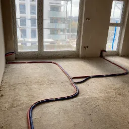 Surowe wnętrze z widocznymi rurami instalacji grzewczej ułożonymi na podłodze, przygotowane do zalania wylewką, z widokiem na budynek w budowie przez okno.