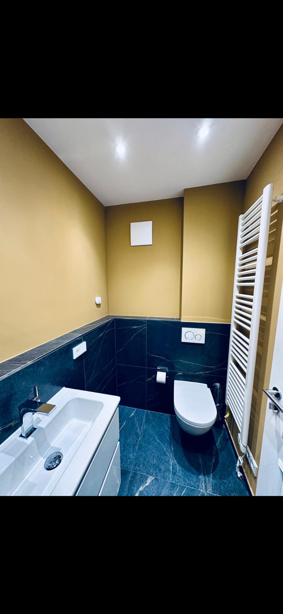 Nowoczesna łazienka z umywalką na szafce, toaletą i grzejnikiem drabinkowym na tle ścian w odcieniach złota i ciemnego marmuru.