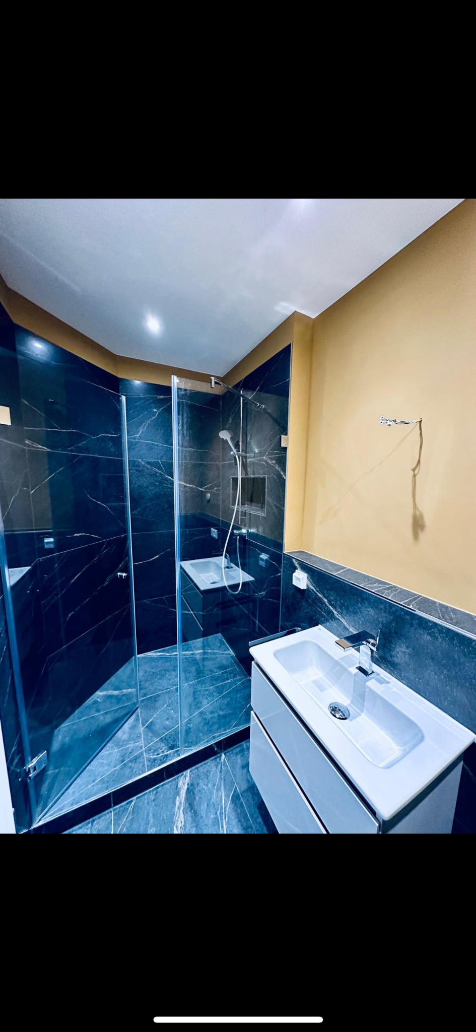 Nowoczesna łazienka z kabiną prysznicową typu walk-in wykończoną czarnym marmurem i białą szafką z umywalką na tle beżowej ściany z wystającymi przewodami.