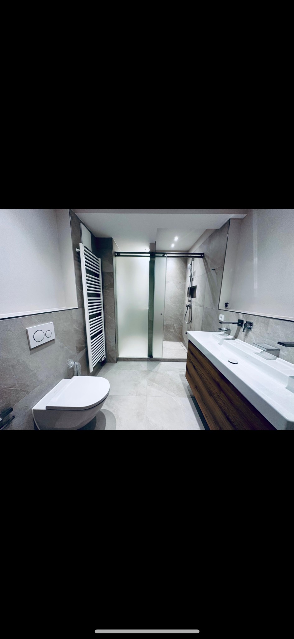 Nowoczesna łazienka z kabiną prysznicową z matowego szkła, wiszącą toaletą, grzejnikiem drabinkowym i drewnianą szafką pod umywalką.