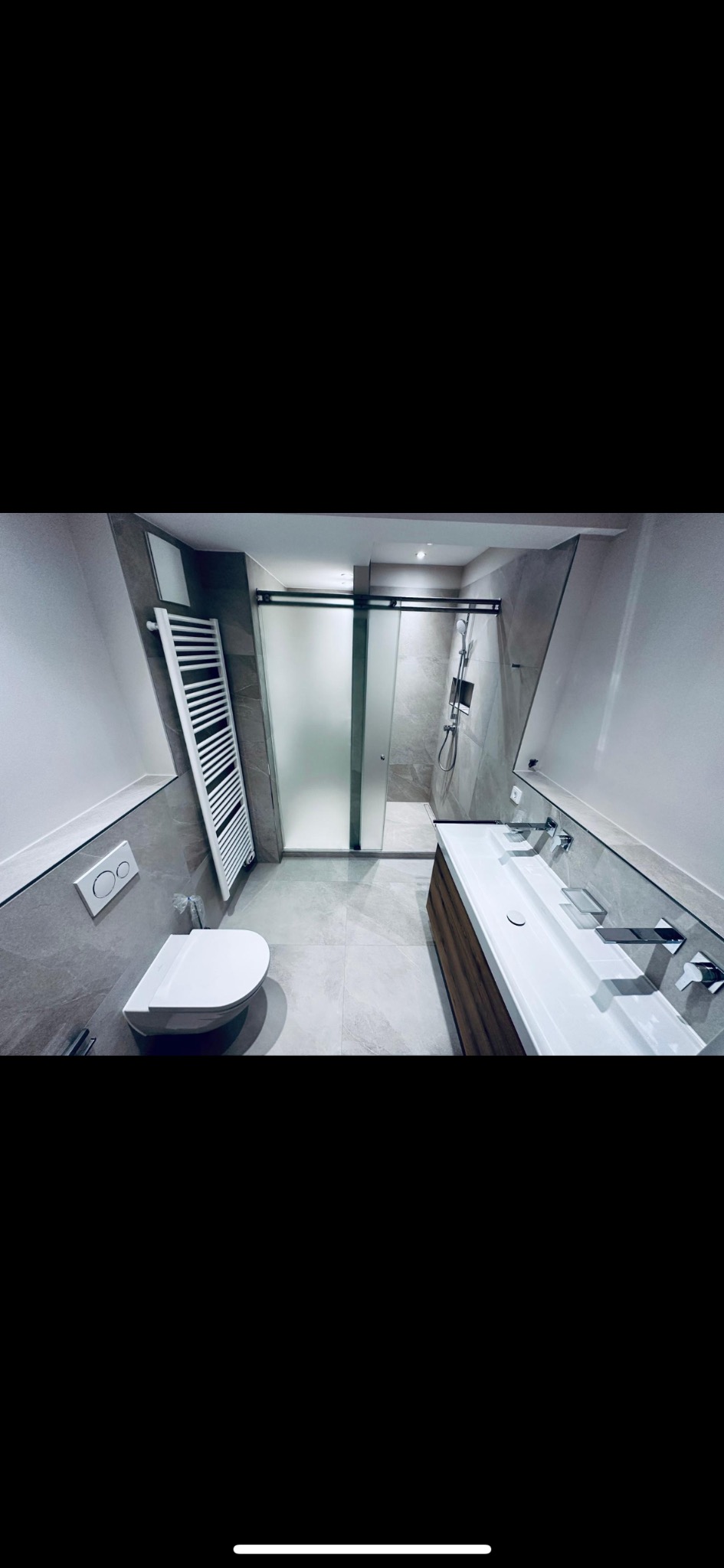 Nowoczesna łazienka na poddaszu z szarą glazurą, kabiną prysznicową ze szkła matowego, wiszącą toaletą, grzejnikiem drabinkowym i podwójną umywalką nablatową z drewnianą szafką.