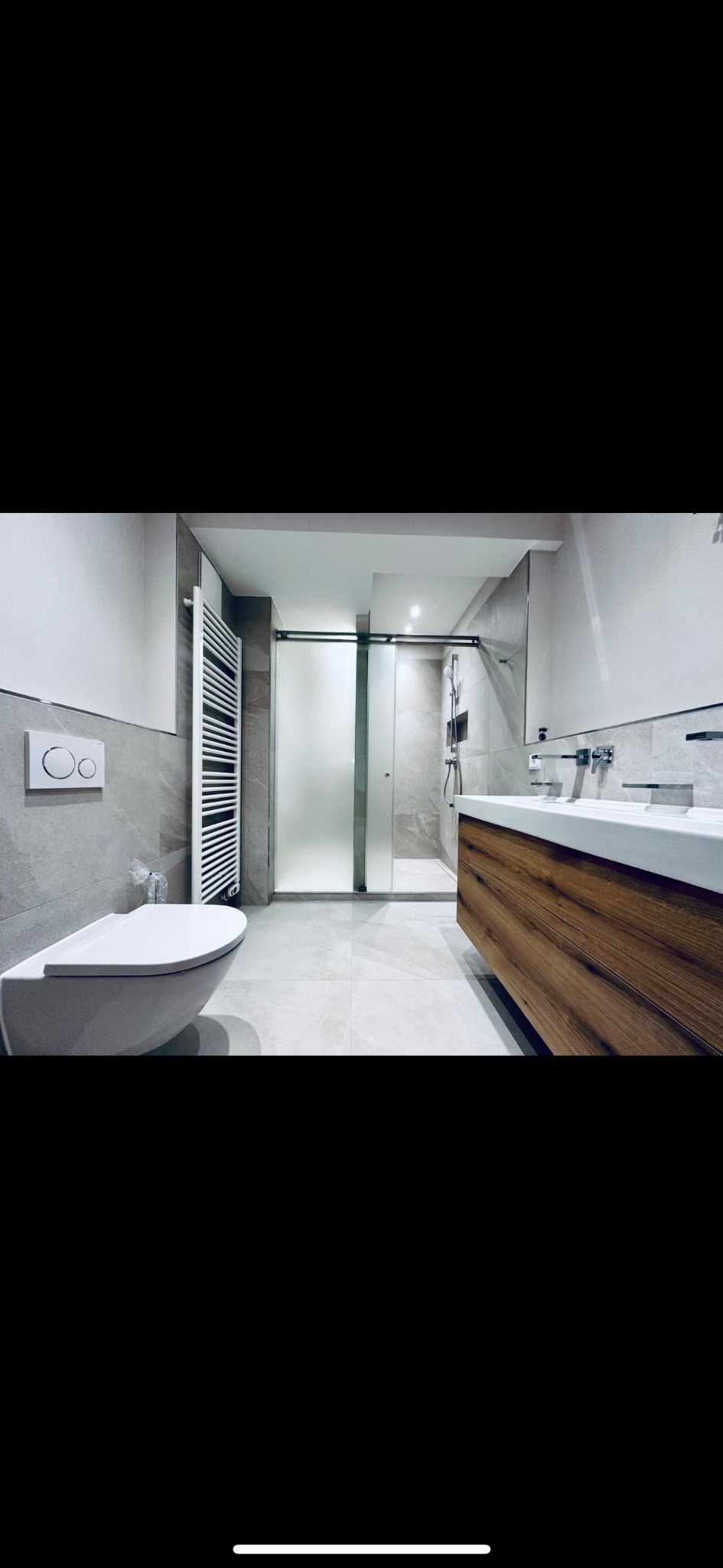 Nowoczesna łazienka z wiszącą toaletą, grzejnikiem drabinkowym, kabiną prysznicową ze szklanymi drzwiami przesuwnymi i umywalką nablatową na drewnianej szafce.