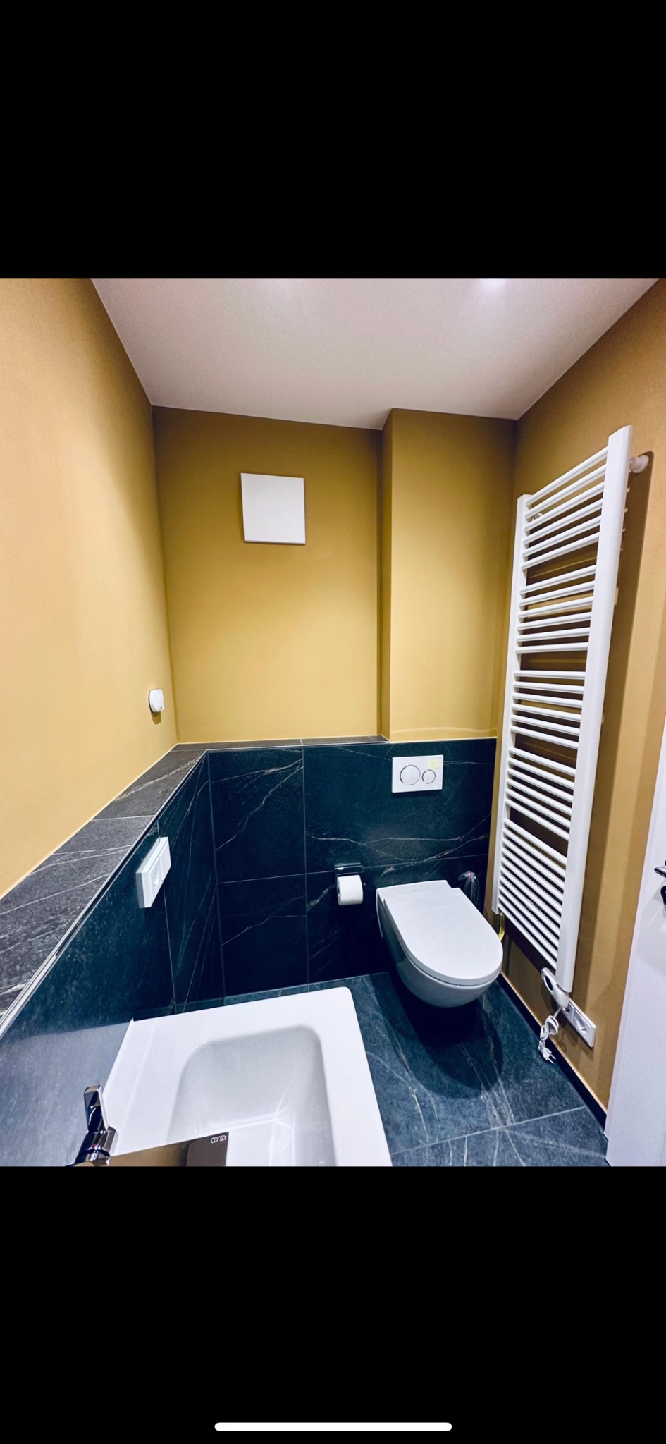 Nowoczesna łazienka z musztardowymi ścianami, czarnymi marmurkowymi płytkami, białą armaturą i grzejnikiem drabinkowym.