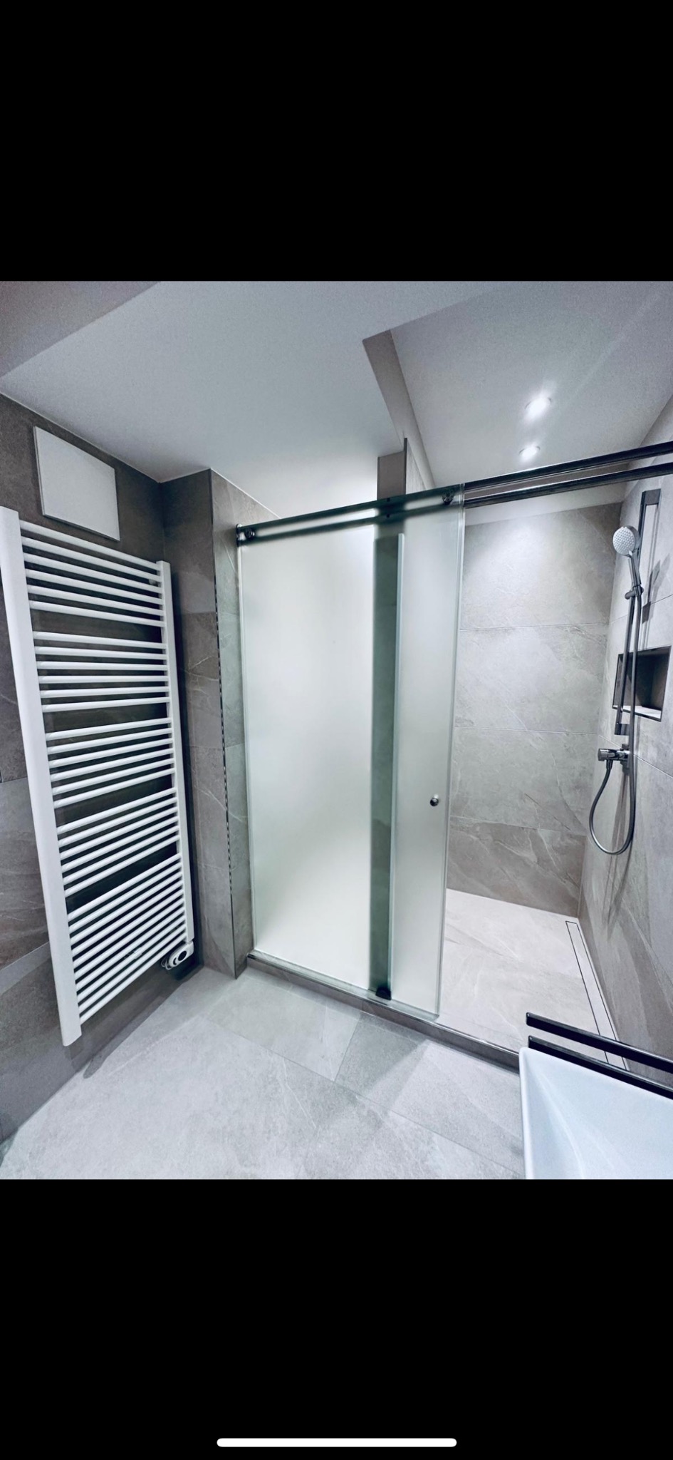 Nowoczesna łazienka z kabiną prysznicową z przesuwnymi drzwiami z matowego szkła, grzejnikiem drabinkowym i szarą płytką imitującą kamień na ścianach i podłodze.