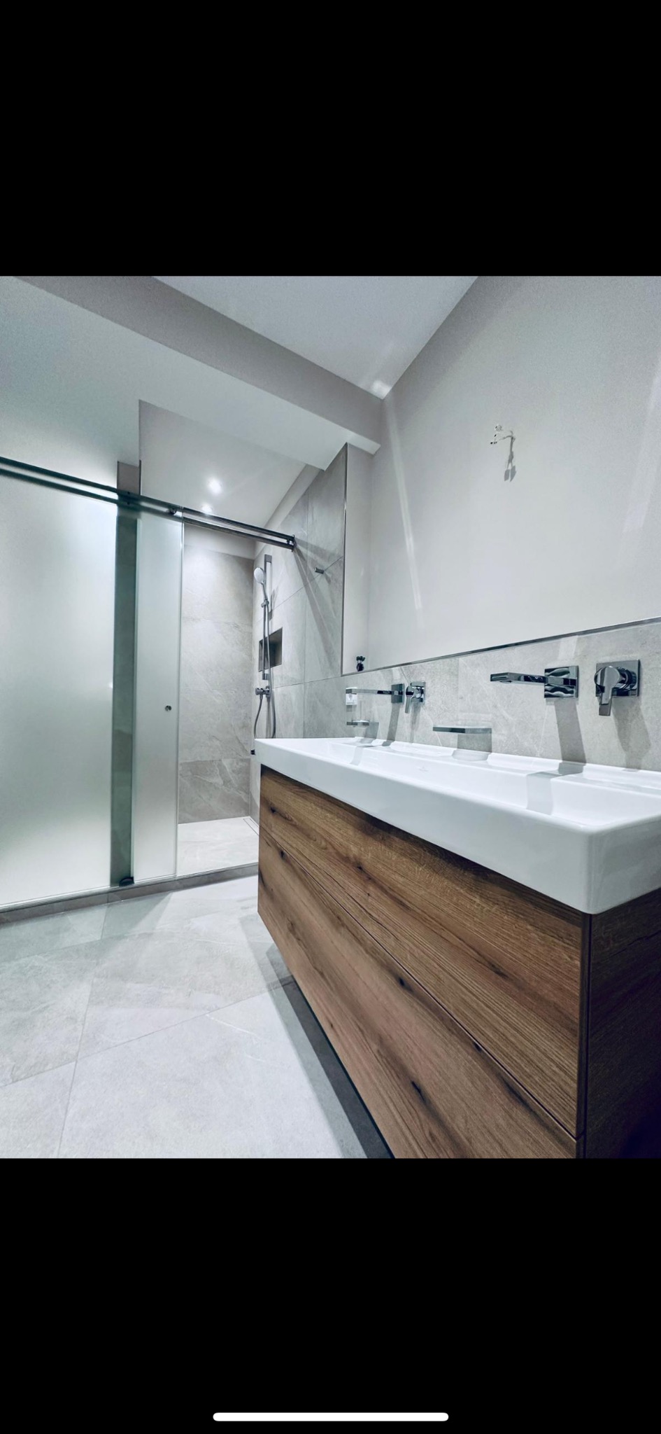 Nowoczesna łazienka z podwójną umywalką nablatową na drewnianej szafce, przeszklona kabina prysznicowa typu walk-in, szara marmurowa płytka na ścianach i podłodze, minimalistyczne baterie.