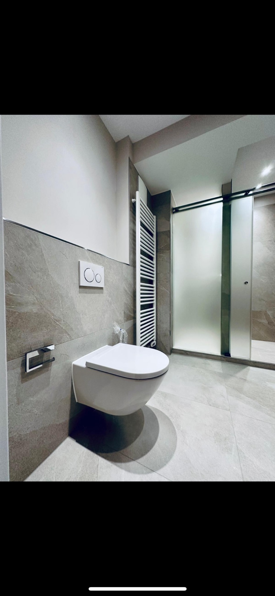 Nowoczesna łazienka z podwieszaną miską WC, grzejnikiem drabinkowym i kabiną prysznicową z przesuwnymi drzwiami, wykończona płytkami w odcieniach szarości.