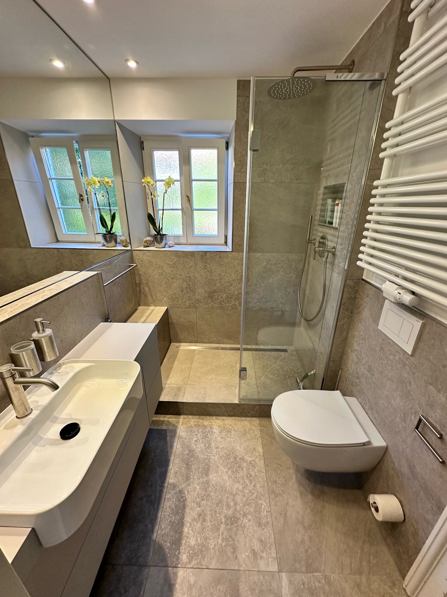 Nowoczesna łazienka z kabiną prysznicową typu walk-in, umywalką nablatową i grzejnikiem drabinkowym, wykończona płytkami w odcieniach szarości z widocznym oknem z kwiatami w doniczkach.