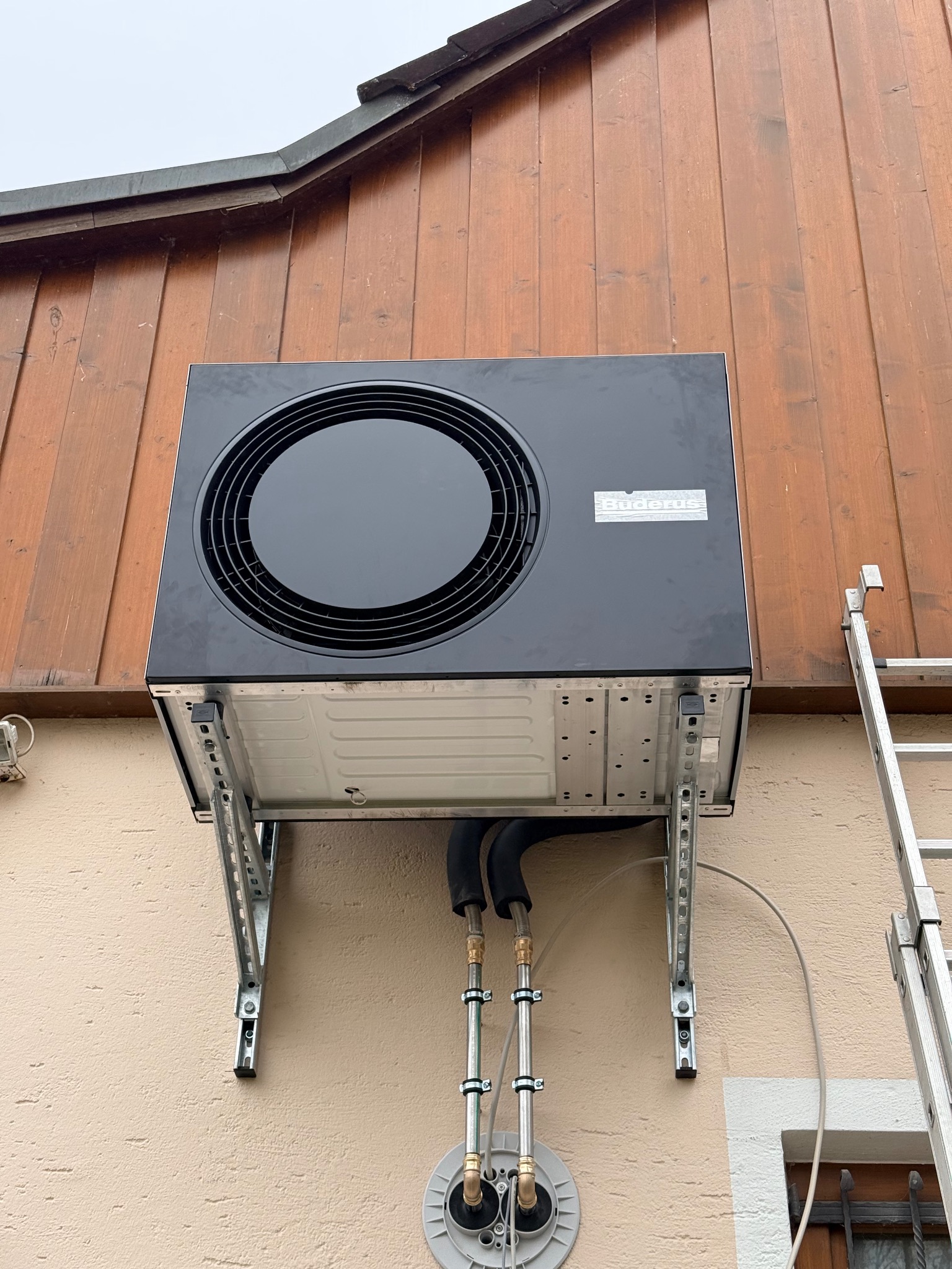 Czarna pompa ciepła Buderus zamontowana na elewacji budynku, widoczne rury i drabina. Ujęcie z dołu, podkreślające solidne mocowanie i estetykę instalacji.