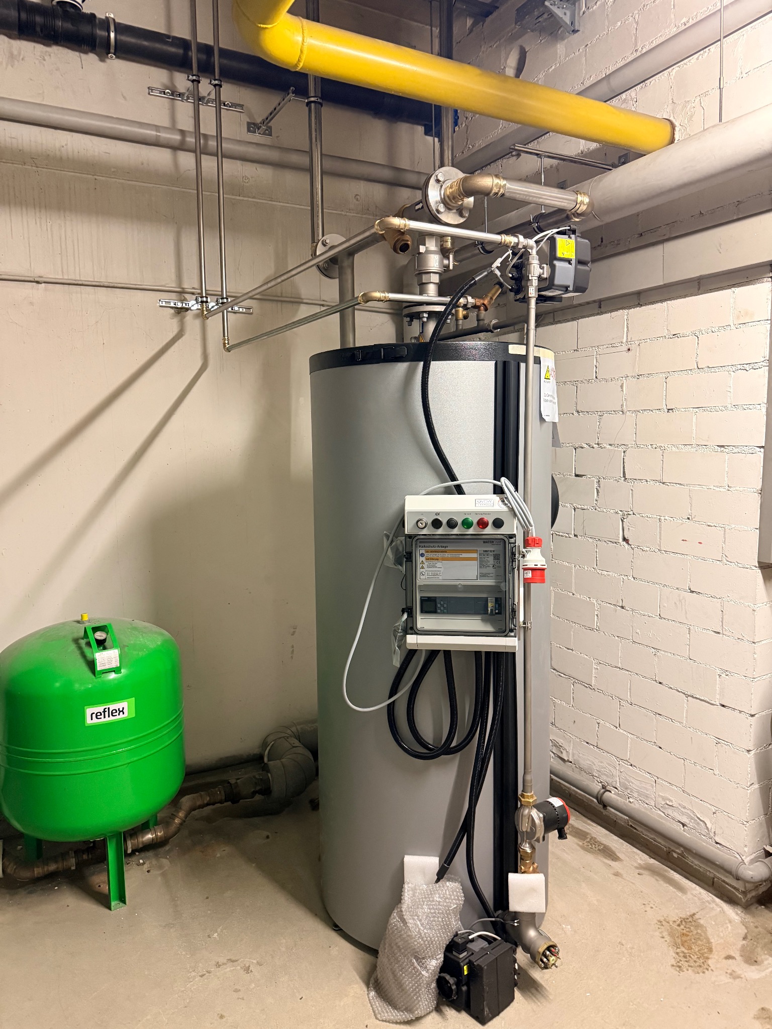Szary bojler z zielonym zbiornikiem Reflex w kotłowni, widoczne rury, instalacja elektryczna i ceglana ściana. Elementy instalacji hydraulicznej.