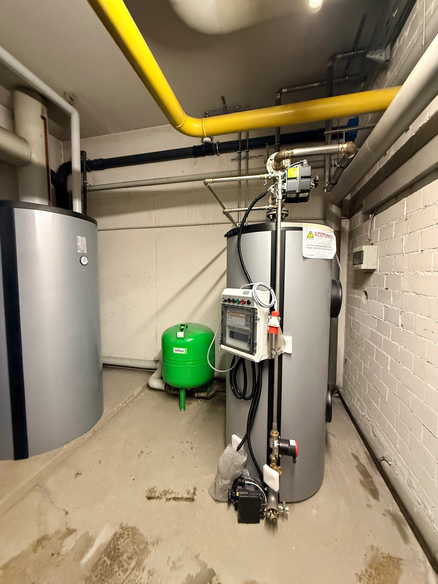 Szary piec z żółtą rurą gazową w kotłowni, obok zielony zbiornik wyrównawczy Reflex. Widoczne rury i instalacje. Ściany z cegły, betonowa podłoga.