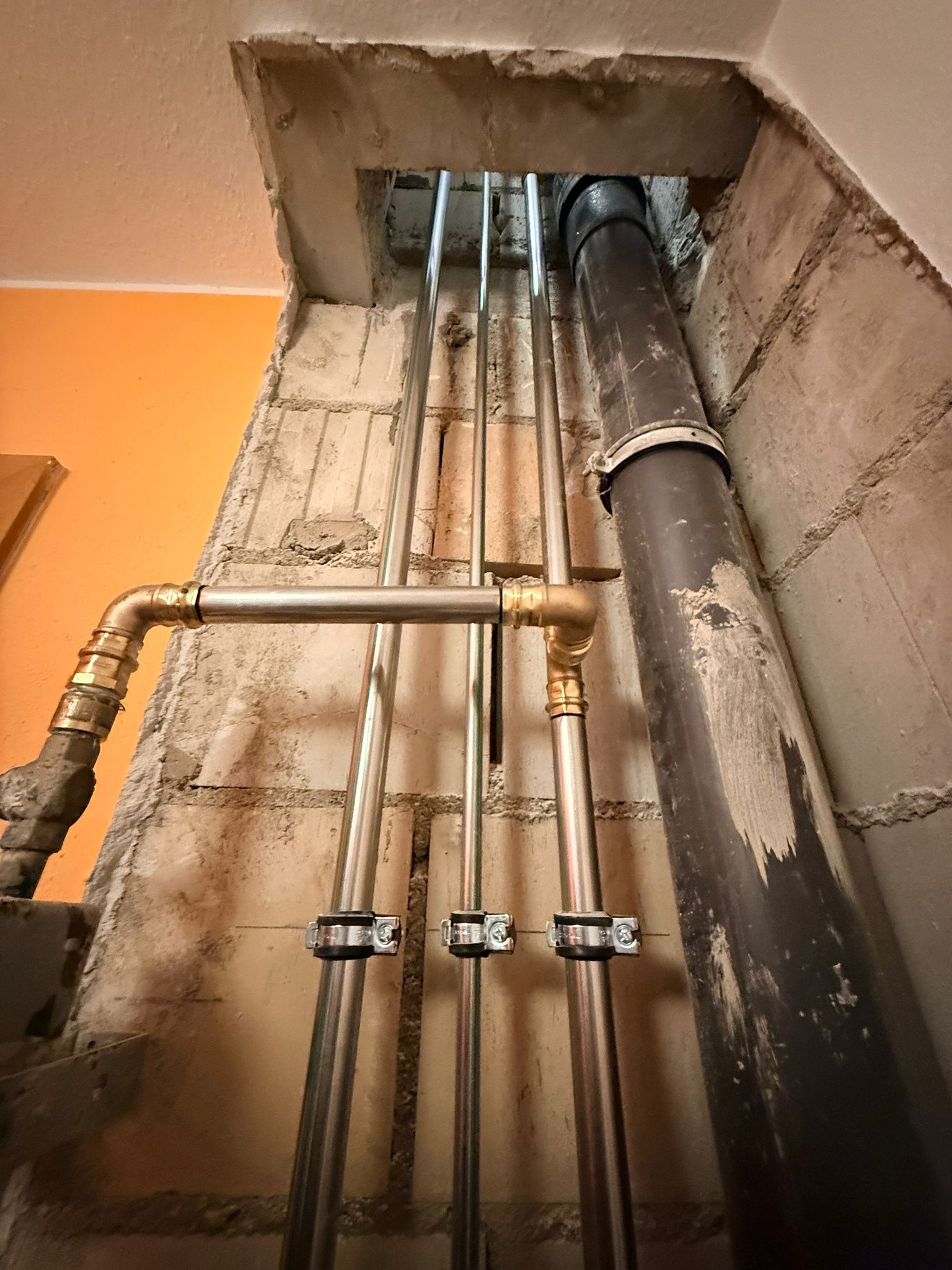 Pionowy układ rur w surowym wnętrzu, z widocznymi mocowaniami i fragmentem instalacji kanalizacyjnej. Ściany z betonu komórkowego, pomarańczowy fragment ściany w tle.