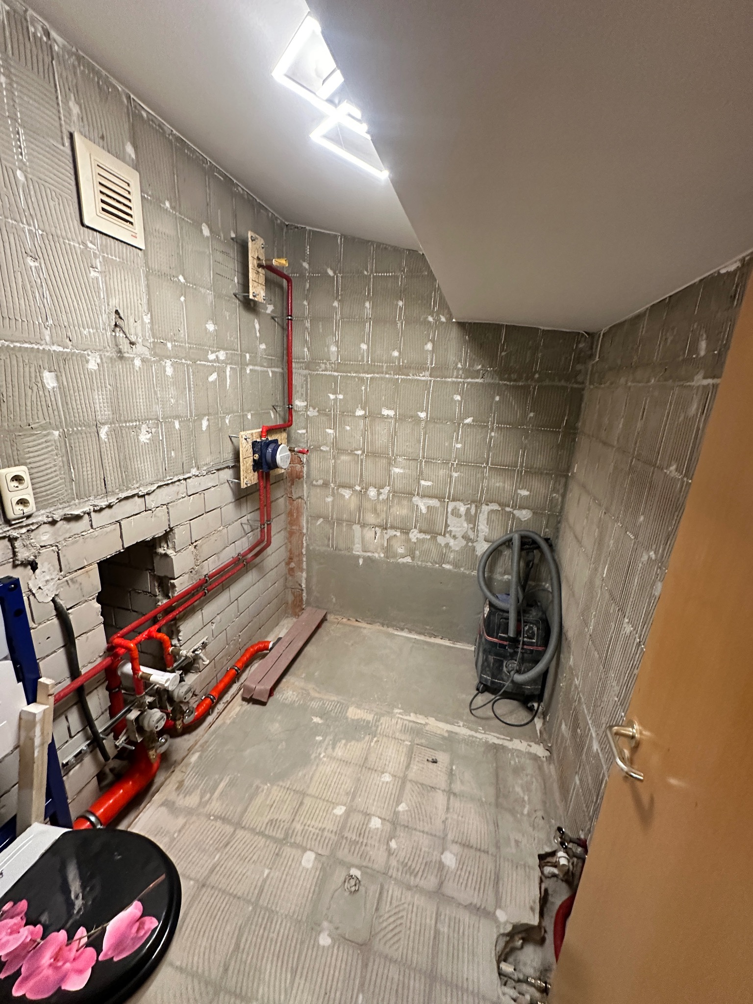 Łazienka w trakcie remontu: widoczne czerwone rury instalacji hydraulicznej na ścianach z resztkami płytek, odkurzacz i deska sedesowa z motywem kwiatowym.