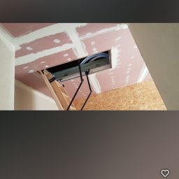 NAT-DOM - Montaż płyt gipsowo-kartonowych na suficie poddasza z widocznym włazem strychowym i schodami rozkładanymi, częściowo wykończone szpachlowaniem.