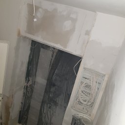 NAT-DOM - Wnętrze w trakcie remontu: widoczne kable elektryczne w puszce podtynkowej, zabezpieczone folią drzwi, surowe ściany z widocznymi śladami gładzi i wystającym przewodem z sufitu.