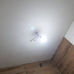 NAT-DOM - Niedokończona instalacja oświetleniowa na suficie w łazience z brązowymi płytkami imitującymi drewno, widoczne kable i prowizoryczna żarówka.