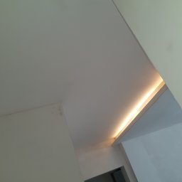 NAT-DOM - Biały sufit z ukrytym oświetleniem LED, widoczne łączenie płyt gipsowo-kartonowych, fragment drzwi w tle.