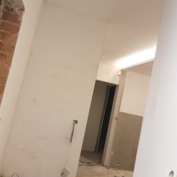 NAT-DOM - Wnętrze w trakcie remontu: świeżo pomalowane ściany na biało, widoczne narzędzia malarskie w rogu, wystające przewody elektryczne i fragment surowej cegły przy framudze.