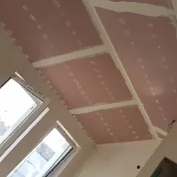 Montaż płyt gipsowo-kartonowych na skośnym suficie poddasza z widocznymi łatami i otworami na punkty oświetleniowe, okna dachowe w tle.