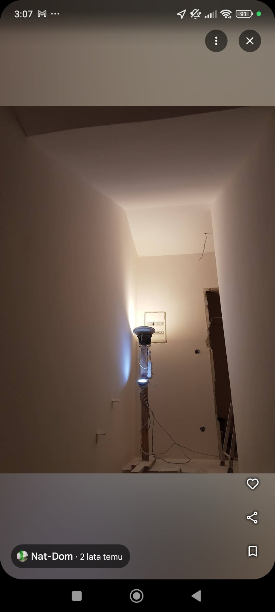 Korytarz w trakcie malowania na biało, widoczna prowizoryczna lampa oświetlająca ścianę, puszka elektryczna i fragment drabiny w przejściu do kolejnego pomieszczenia.